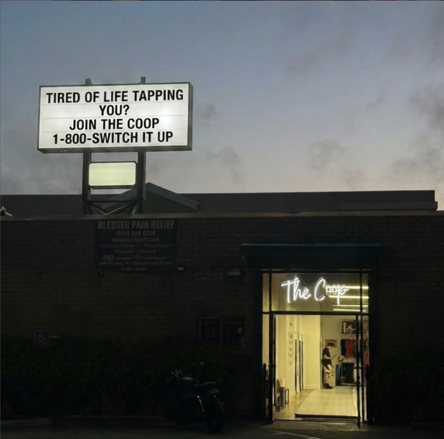 The Coop storefront at dusk — neon script glowing in the doorway, the roadside letterboard reading 'Tired of life tapping you? Join The Coop.'